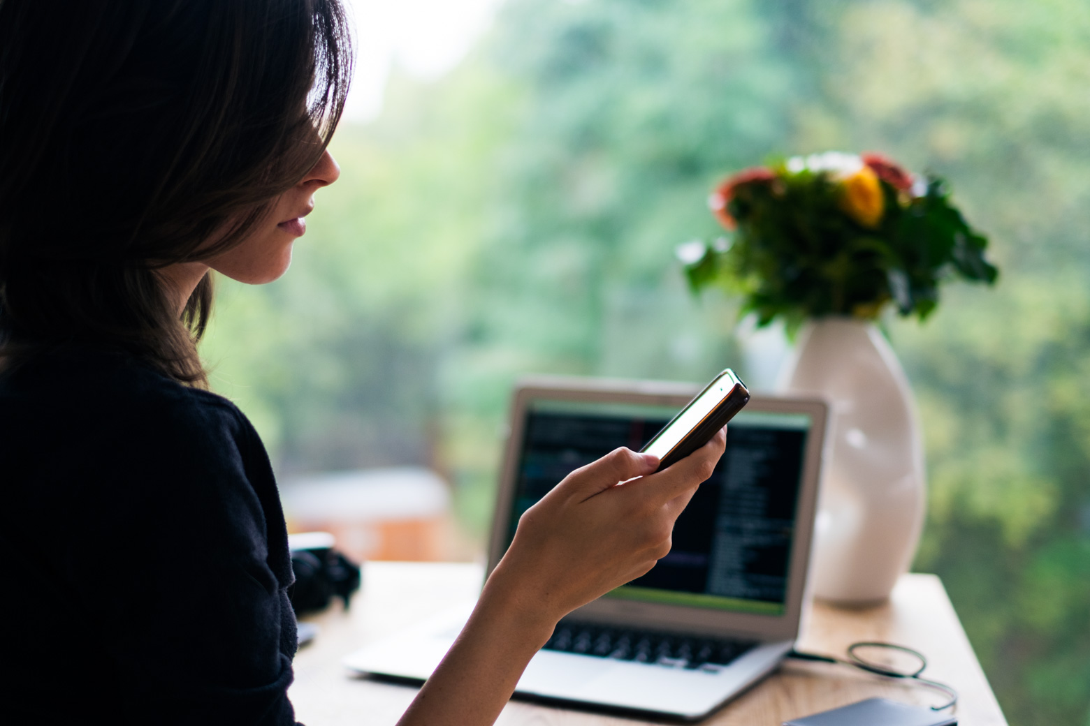 woman holding mobile phone and laptop with flower vase on the table