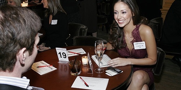 a smiling woman sitting crossed legs in front of a man and a round table between them with a water glass, candle, papers and pens on top of it