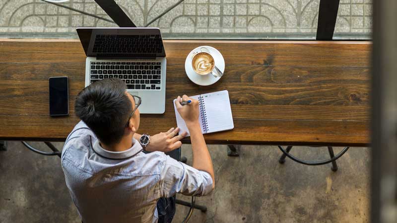 man writing in the notebook with laptop in a wooden table at a coffee shop