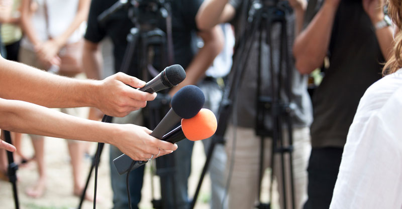 two hands holding microphones and media people around