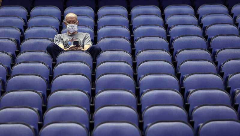 a man with a face mask while looking at his phone sits in a blue chair alone in an empty Coliseum