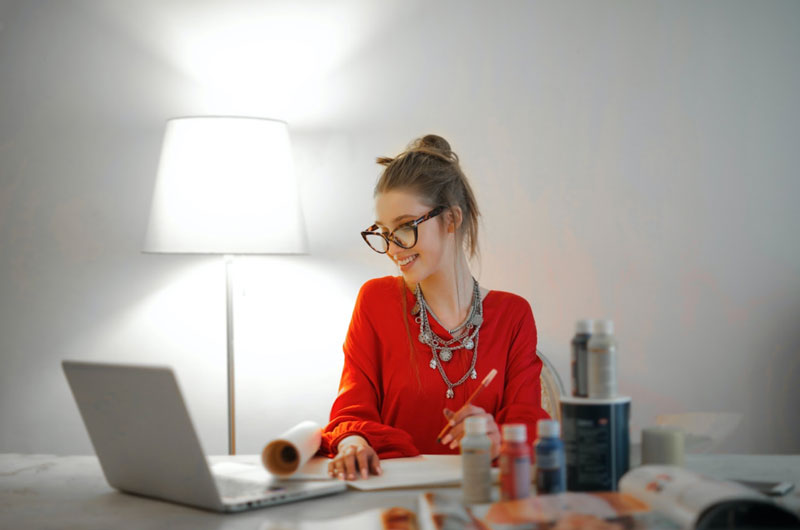 a smiling woman wearing eyeglasses and red top looks at laptop computer